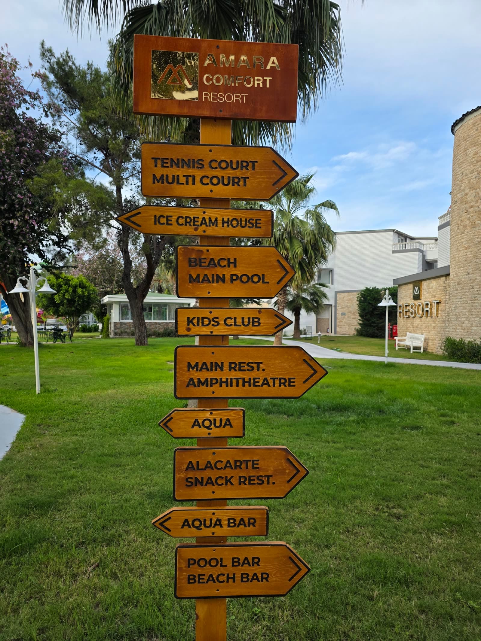 Indicator de lemn în grădina hotelului, cu direcțiile către terenul de tenis, plajă și piscina principală, kids club, restaurantul principal, amfiteatru, zona aqua și barurile de la piscină și plajă.