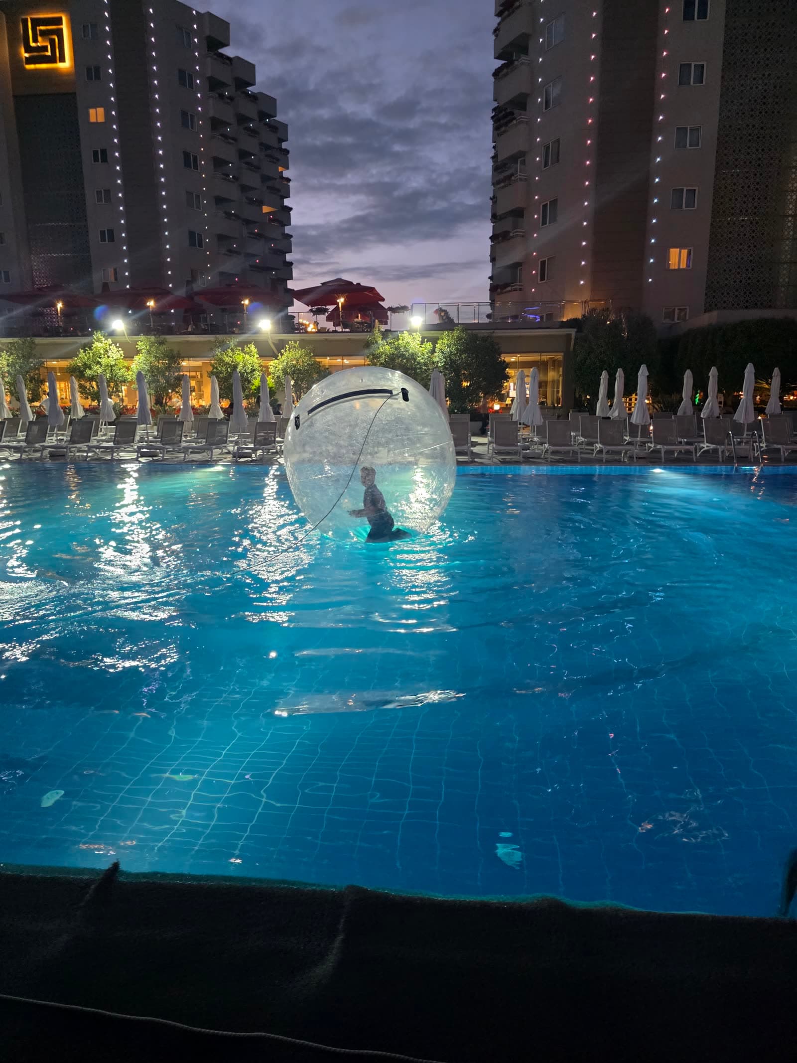 Piscina hotelului seara, cu o bilă gonflabilă transparentă pe apă și șezlonguri în jur, sub luminile terasei.