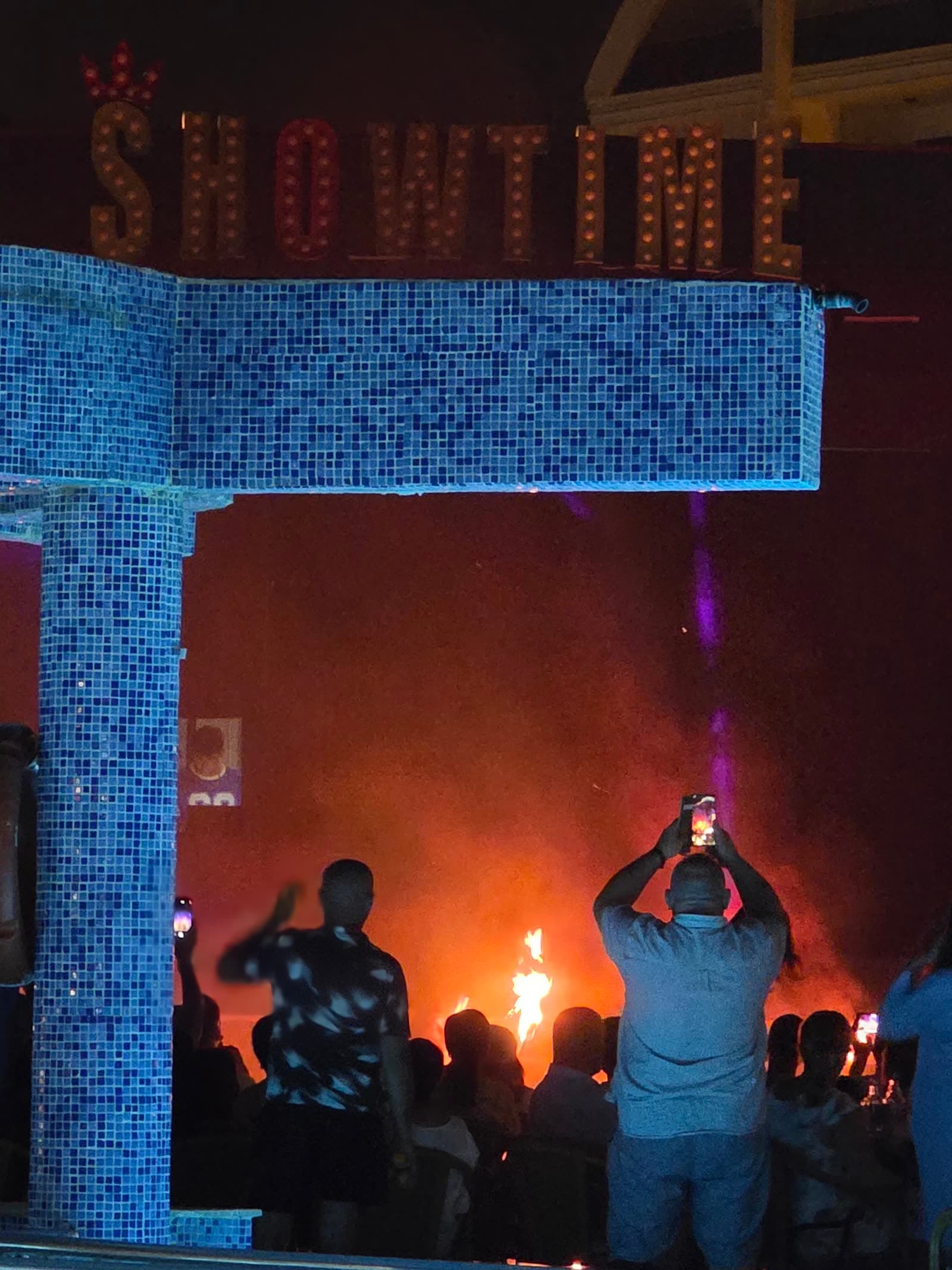 Scena hotelului noaptea, cu flăcări în fundal și spectatori care filmează cu telefoanele, sub firma luminoasă „Showtime”.