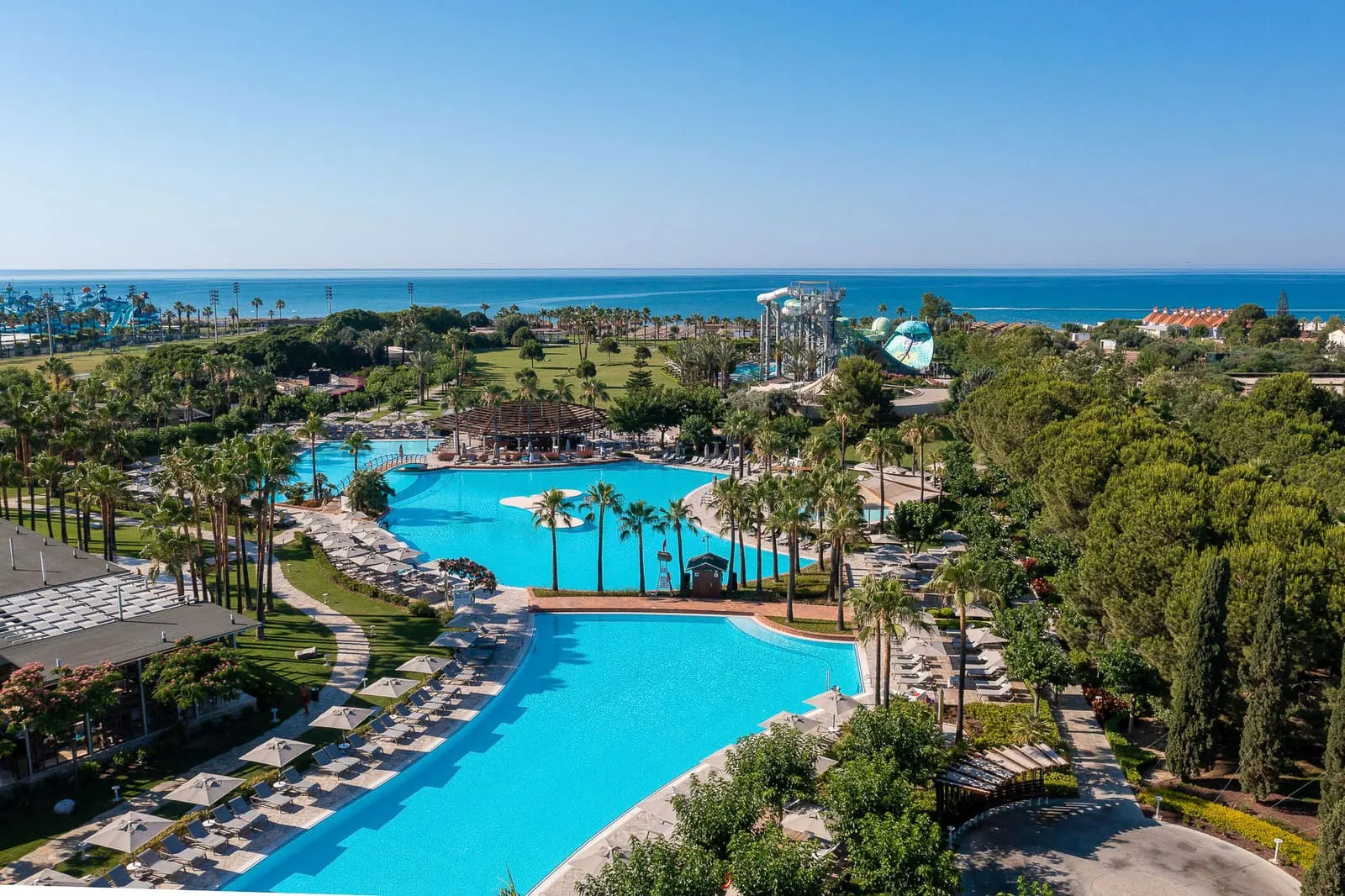Piscinele înșirate spre mare, cu șezlonguri și umbrele pe margini și toboganele aqua park-ului în depărtare.