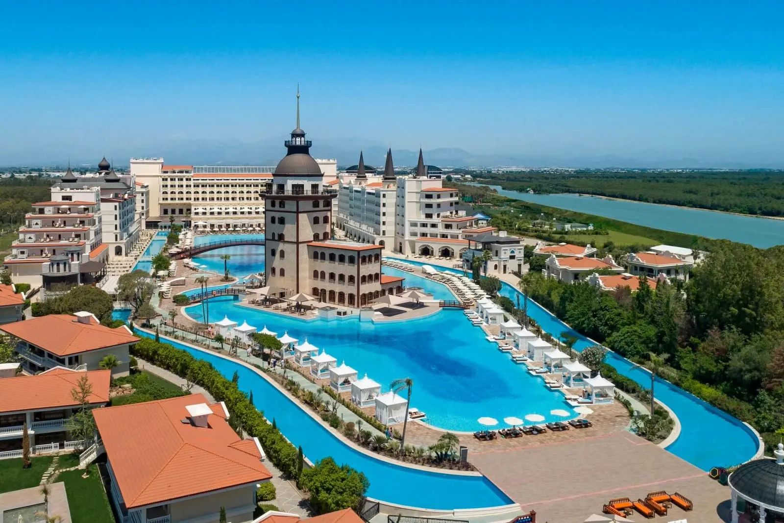 Vederea aeriană de zi: turnul cu cupolă în mijlocul piscinei care șerpuiește printre clădiri, cu râul și pădurea alături.