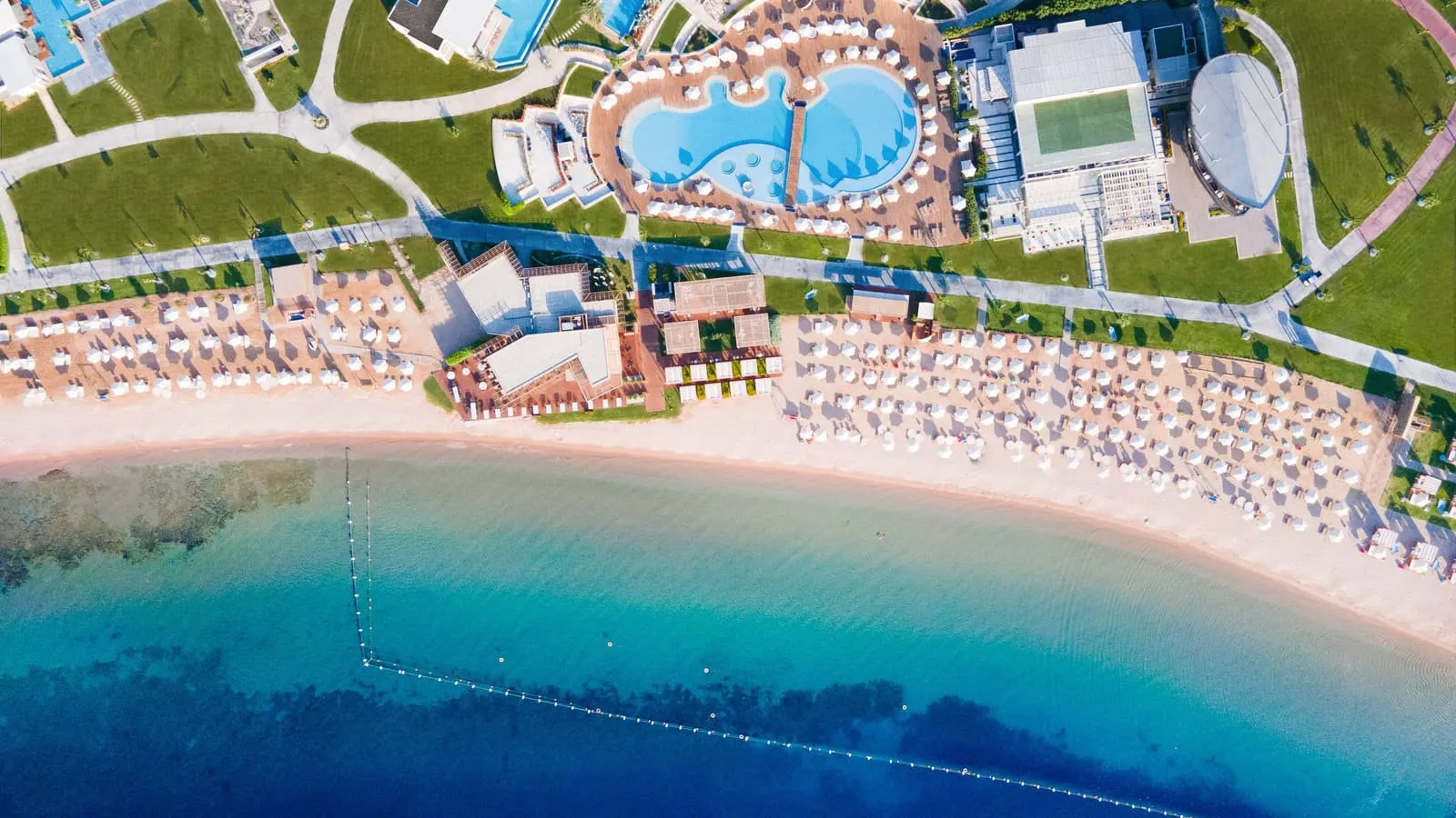 Plaja văzută de deasupra: rânduri de baldachine albe pe nisip, apa limpede cu recif și piscina în formă de paletă între clădiri.