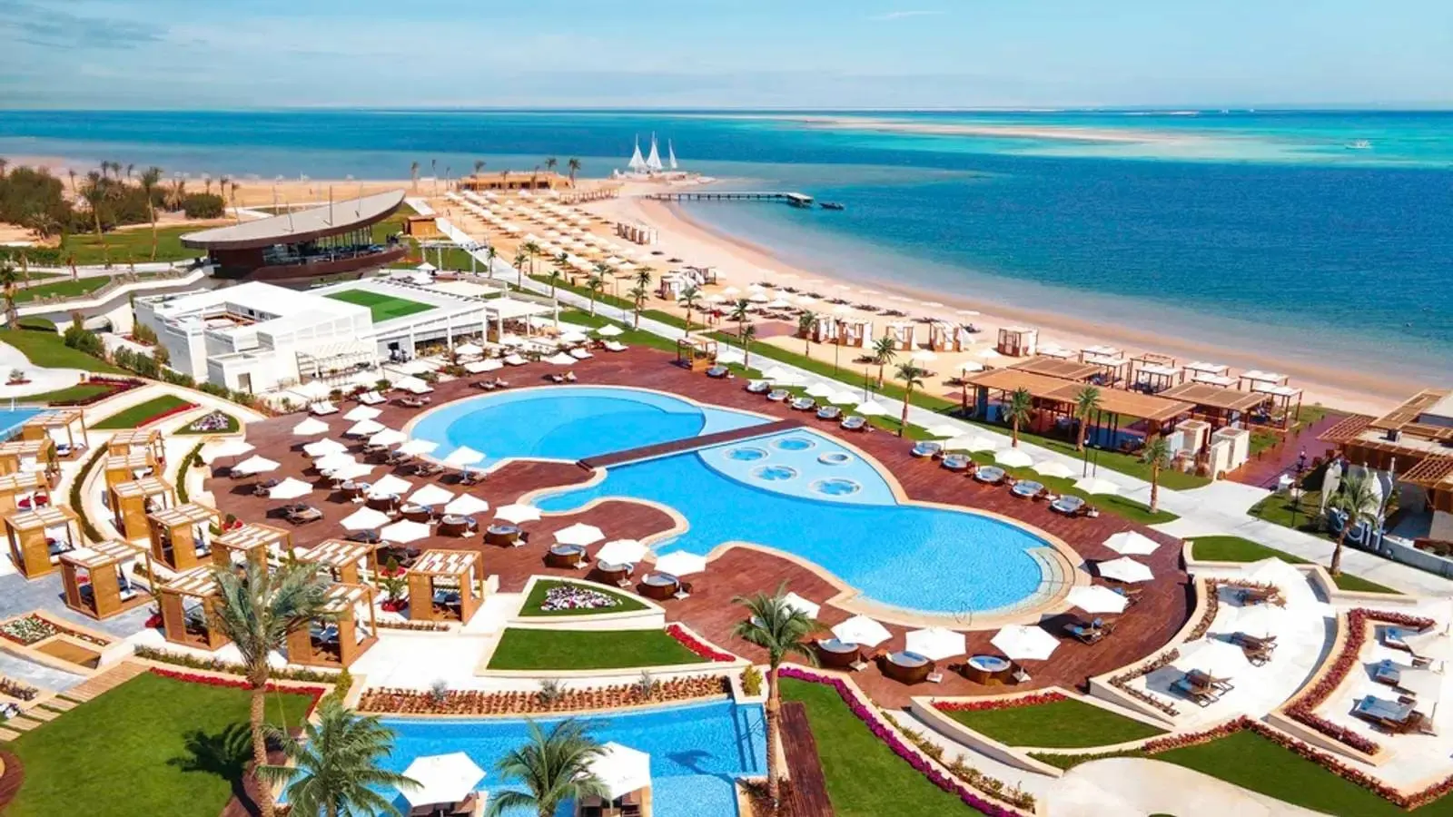Stațiunea văzută de sus: piscinele curbate cu jacuzzi-uri rotunde, plaja lungă de nisip cu șiruri de umbrele, pontonul și bancurile de nisip turcoaz ale Mării Roșii.